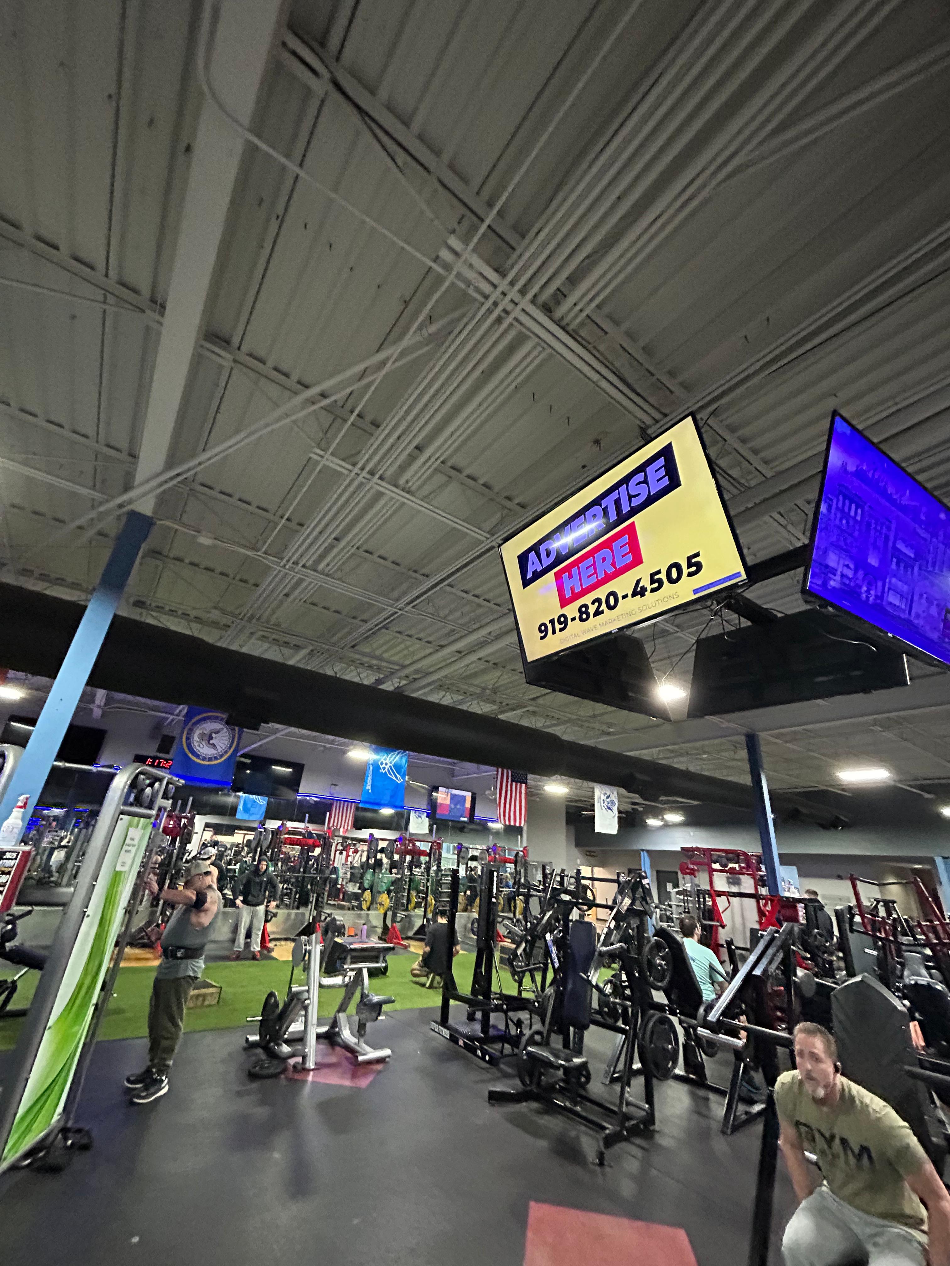 Digital billboard suspended above gym equipment in a fitness venue